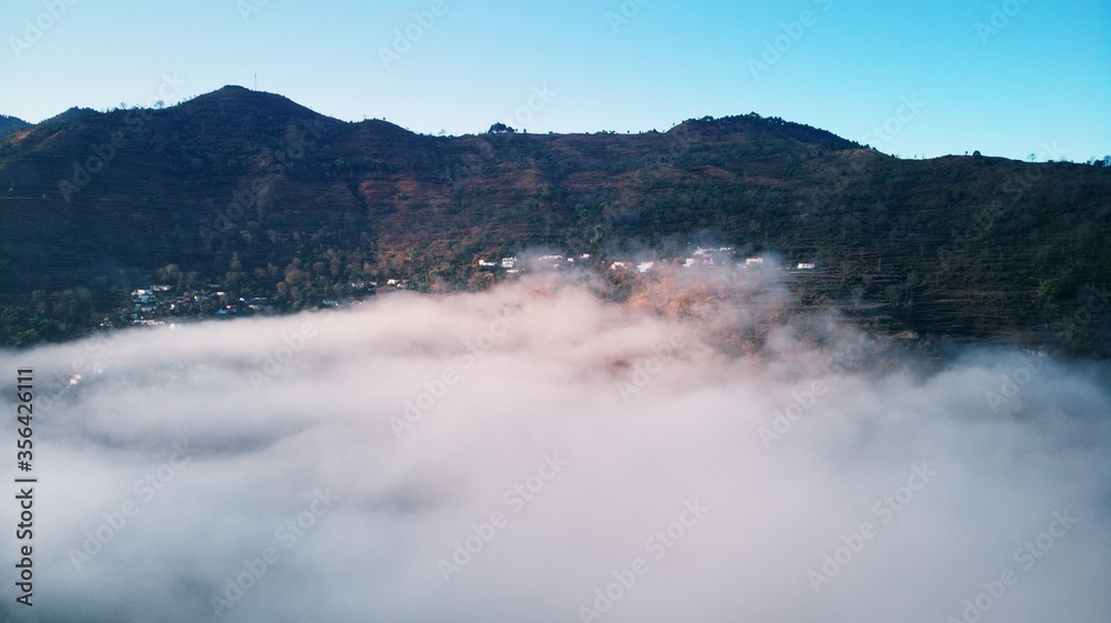 Aerial/Drone View of a foggy Himalayan valley and green fields surrounded by lush green hills during a beautiful morning at a rural location in Uttarakhand, India. Aerial/Drone View of a foggy Himalayan valley and green fields surrounded by lush green hills during a beautiful morning at a rural location in Uttarakhand, India.