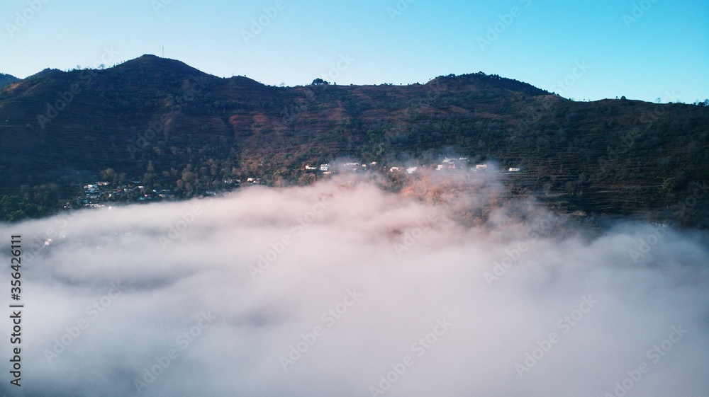 Aerial/Drone View of a foggy Himalayan valley and green fields surrounded by lush green hills during a beautiful morning at a rural location in Uttarakhand, India.