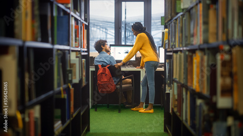 University Library: Boy Uses Personal Computer at His Desk, Talks with Girl Classmate who Explains, Helps Him with Class Assignment. Focused Students Study Together. Shot Between Rows of Bookshelves