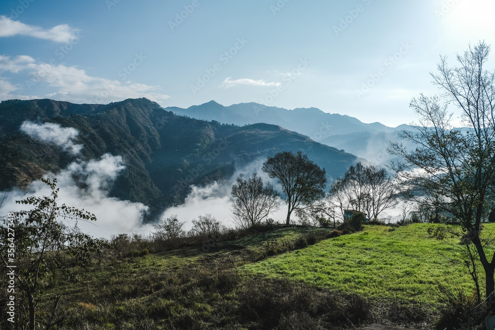 Aerial/Drone View of a foggy Himalayan valley and green fields surrounded by lush green hills during a beautiful morning at a rural location in Uttarakhand, India.