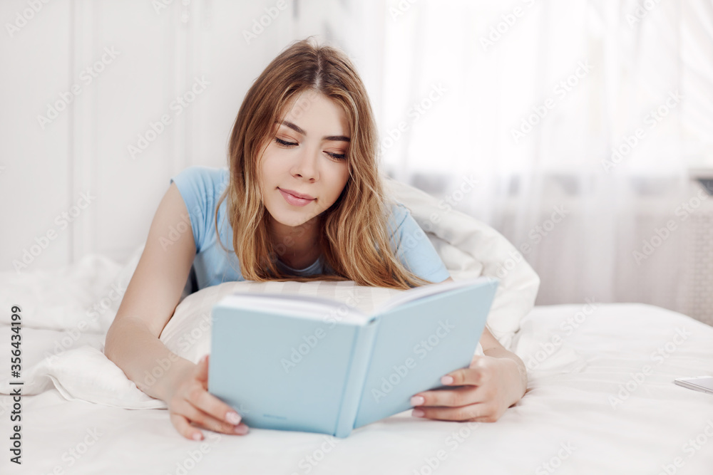 Beautiful girl in blue pajamas reading a book, lying on the bed in the bedroom. Training, education, Hobbies. Beautiful girl in blue pajamas reading a book, lying on the bed in the bedroom. Training, education, Hobbies.
