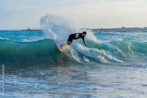 Spanish young man practicing skimboard on Mallorca's beaches with blue waves and clear sky with a black and blue wetsuit