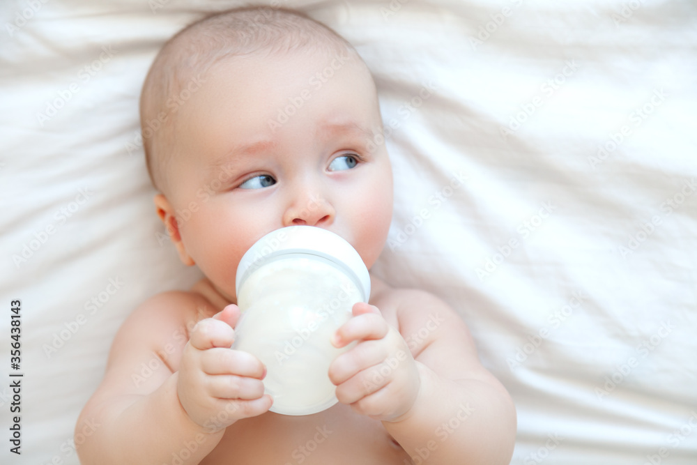 Beautiful baby eating milk from bottle in white bedroom on white background Beautiful baby eating milk from bottle in white bedroom on white background