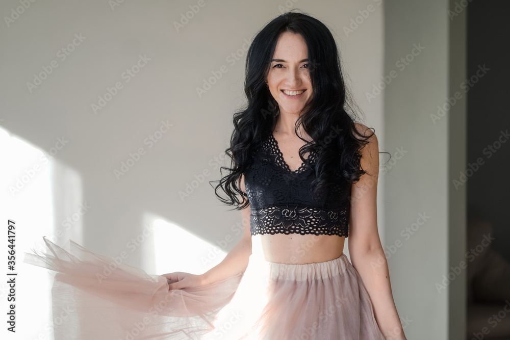 Fototapeta premium beautiful happy girl in a black top and pink skirt against the gray walls near the window