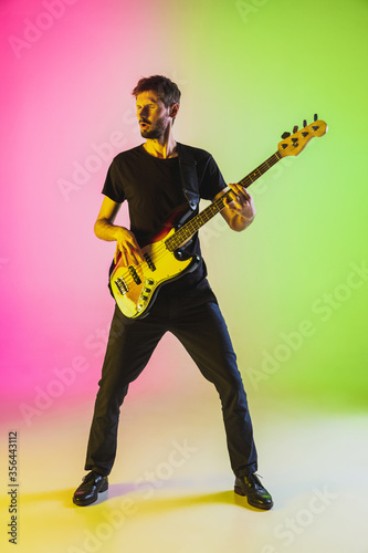 Young and joyful caucasian musician playing bass guitar on gradient studio background in neon light. Concept of music, hobby, festival. Colorful portrait of modern artist. Inspired improvising.