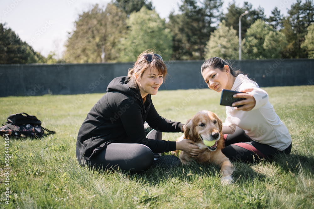 Smiling women sitting in park with dog and taking selfie. Pretty girls taking selfie while sitting on meadow with retriever dog and ball. Smiling women sitting in park with dog and taking selfie. Pretty girls taking selfie while sitting on meadow with retriever dog and ball.