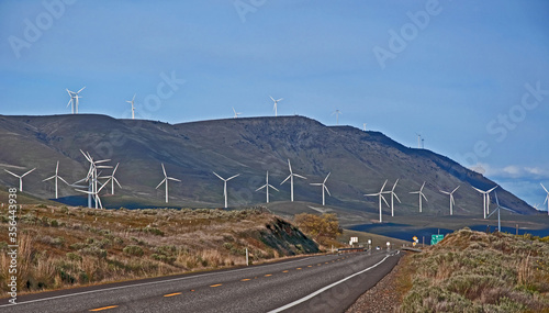 This landscape image shows many modern white tall windmill power generators in action turning their propellers along a rural highway with dry, arid mountains and blue sky in the photo. Taken in the 