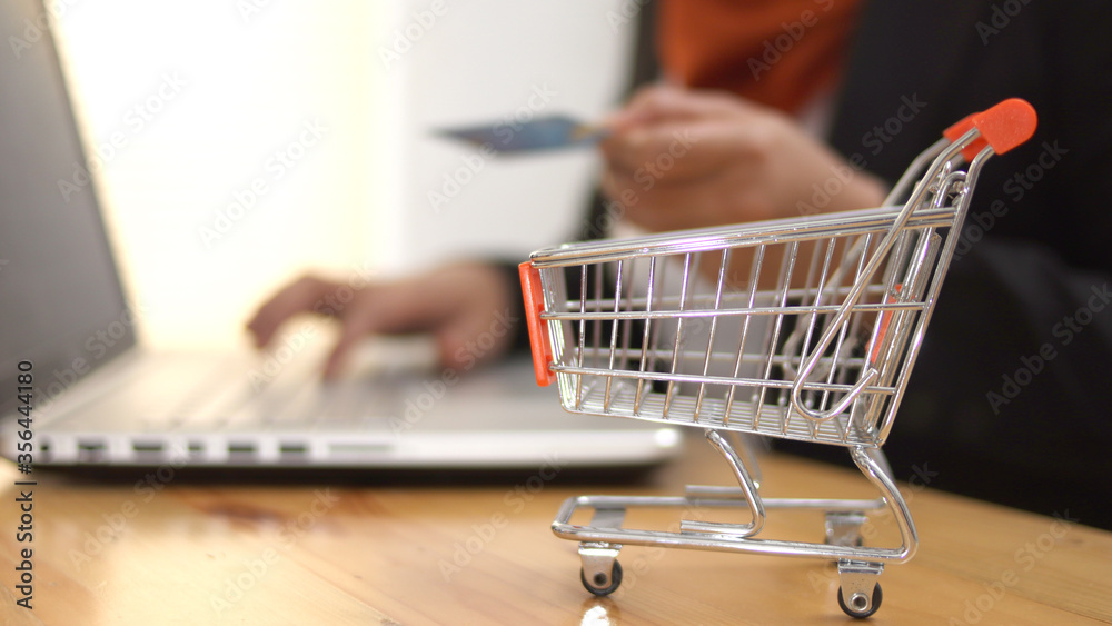 Shopping online on internet store, close up of mini shopping cart trolley with woman making purchase on laptop with credit card in the background