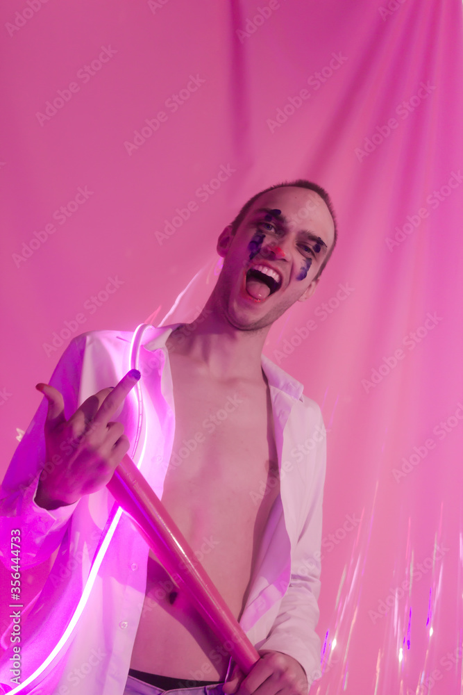Portrait of Young Handsome emotional man in an unbuttoned white shirt with pink bat. Male in the image of a crazy clown like a joker. Neon pink room. Reflect of neon. Selective focus. Portrait of Young Handsome emotional man in an unbuttoned white shirt with pink bat. Male in the image of a crazy clown like a joker. Neon pink room. Reflect of neon. Selective focus.
