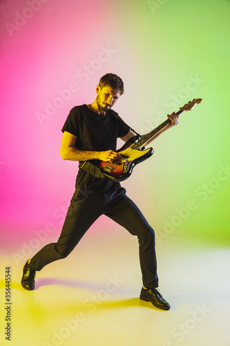 Close up young caucasian musician playing bass guitar on gradient studio background in neon. Concept of music, hobby, festival. Colorful portrait of modern artist. Inspired, impressive improvising.