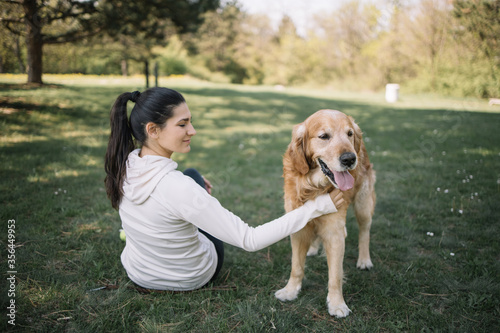 Brunette woman with eyes closed petting dog in nature. Dog standing on meadow and woman petting him while sitting on ground in forest.