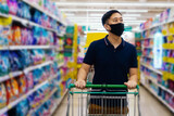 Young adult Asian man wearing a protective mask while shopping with cart trolley in grocery supermarket store. He's choosing to buy products in the market during Covid 19 crisis.