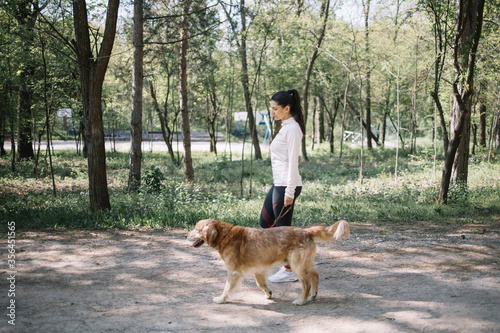 Side view of girl and dog having a walk in park. Brunette woman in sport outfit walking on park alley with dog.