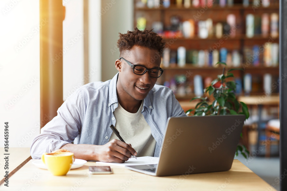 Obraz premium Millennial African American student with laptop taking notes during breakfast in cafe