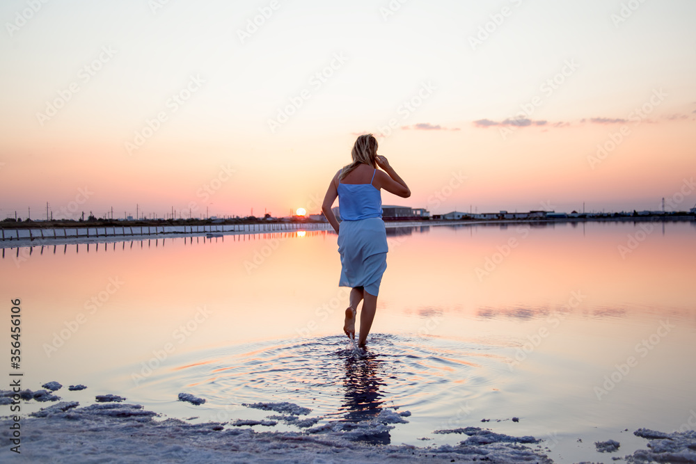 Fototapeta premium Rear view of the woman back look to the purple sunrise in water on summer. Woman walking on the shore of the salt beach at pink lake. vacation lifestyle near ocean