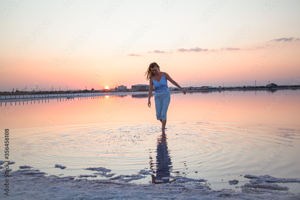 Fototapeta premium Beautiful sensual woman walking and look to the purple sunrise in water on summer. Woman sit on the shore of the salt beach at pink lake. vacation lifestyle near ocean
