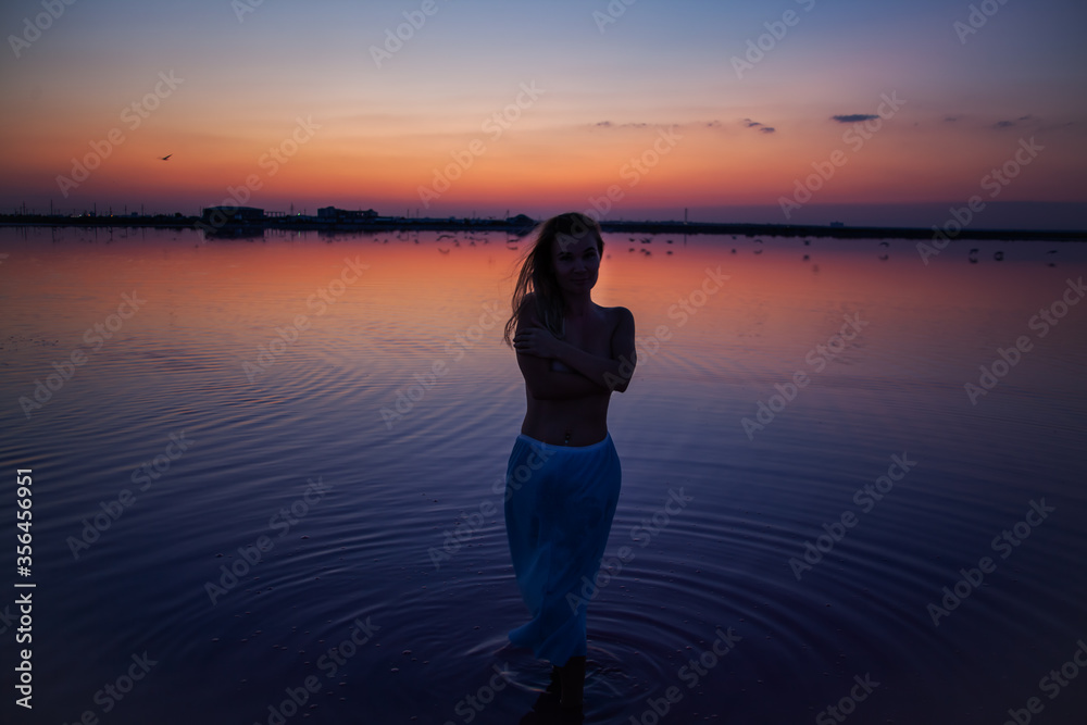 Fototapeta premium The silhouette of woman look to the purple sunrise in water on summer. Woman sit on the shore of the salt beach at pink lake. vacation lifestyle near ocean