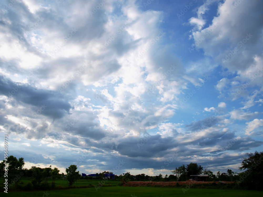 White gray clouds in the blue sky reflect the beautiful evening light. Below is a green field and trees. Natural beauty in the rainy season. Selective focus