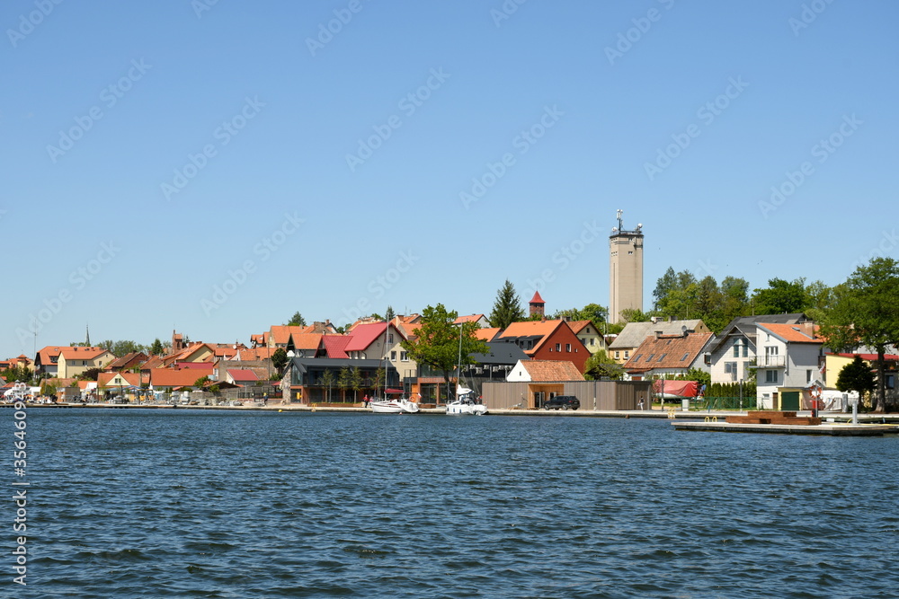 A view of a small port city or town in Poland with a big water tower overlooking small buildings and hotels located next to the coast of a vast yet shallow river seen on a cloudless summer day