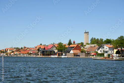 A view of a small port city or town in Poland with a big water tower overlooking small buildings and hotels located next to the coast of a vast yet shallow river seen on a cloudless summer day