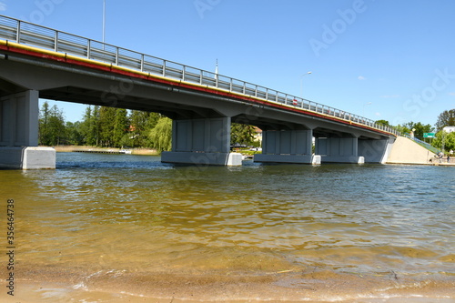 A view of a pedestrian bridge made out of concrete and decorated with metal and wood elements with a vast yet shallow lake or river flowing below seen on a sunny spring day in Poland
