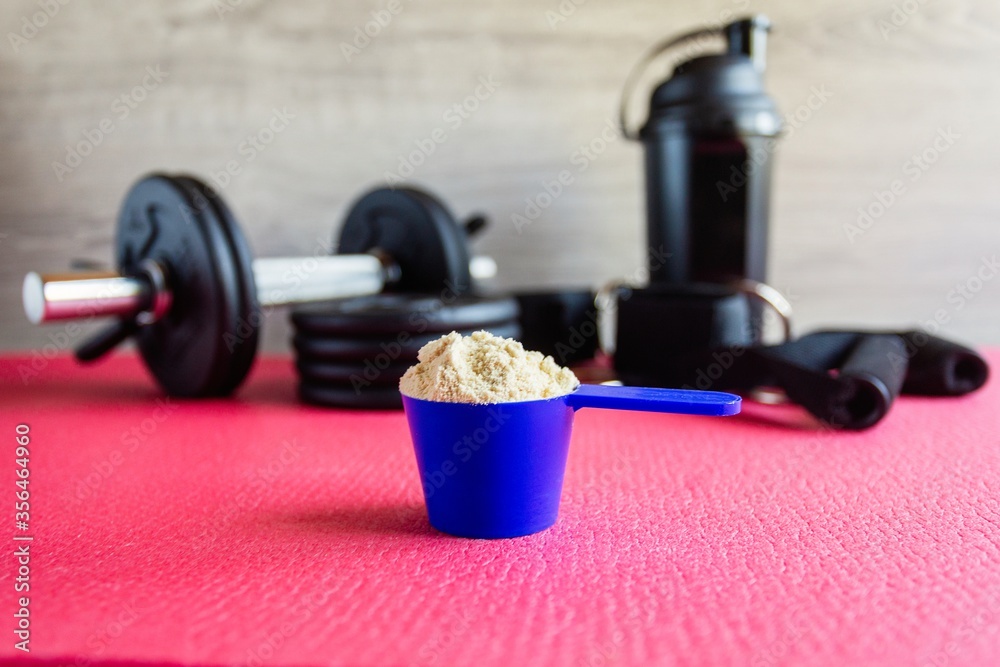 Plastic spoon or measuring scoop of whey protein on the background of sports equipment, a shaker for mixing the mixture, black dumbbells, leg ties, elastic bands for traction and attachment handles. Plastic spoon or measuring scoop of whey protein on the background of sports equipment, a shaker for mixing the mixture, black dumbbells, leg ties, elastic bands for traction and attachment handles.