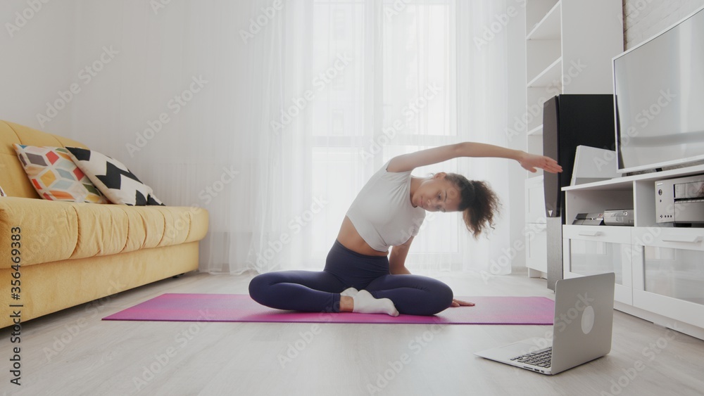 On-line home work out woman using internet services with help of her instructor on laptop at home. Slim sporty african american woman practicing yoga on a mat. Woman stretching back