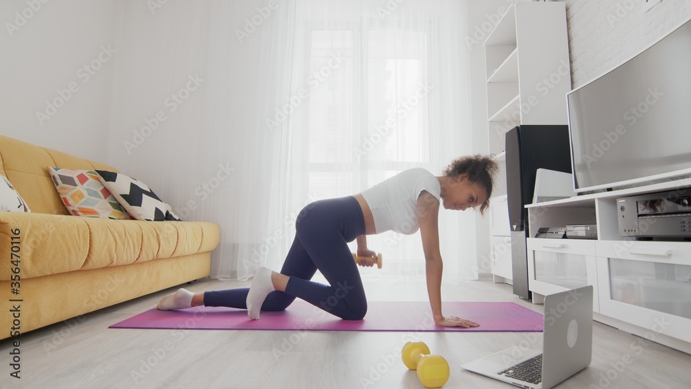 On-line home work out woman using internet services with help of her instructor on laptop at home. Slim sporty african american woman pulling up a yellow dumbbell on mat On-line home work out woman using internet services with help of her instructor on laptop at home. Slim sporty african american woman pulling up a yellow dumbbell on mat