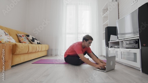 Androgynous Transgender beautiful young gay man with make up making sits on mat and uses the laptop. On-line home work out man using internet services with help of her instructor on laptop at home.