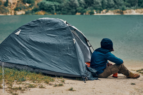 Young man sitting by the tent in nature and looking at beautiful lake. Weekend camping concept.