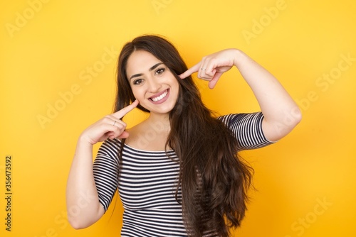 Photo of happy young woman smiling and pointing her cheeks isolated over yellow background