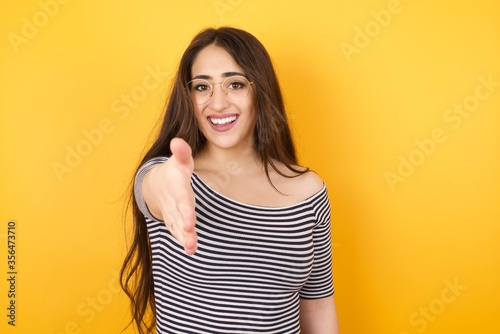 Portrait of cheerful woman shake hand greet person wearing glasses and stripped T-shirt, standing against yellow background. Greetings and welcoming concept.