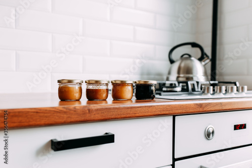 Little honey cans on the wooden table. Honey variety cans in a row. Bright kitchen interior. Wooden complete kitchen with gas oven