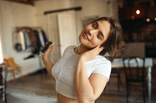 Beautiful young woman in tight fitting cropped top having relaxed carefree facial expression keeping eyes closed  enjoying nice tracks in earphones. Music  dancing  leisure and relaxation concept