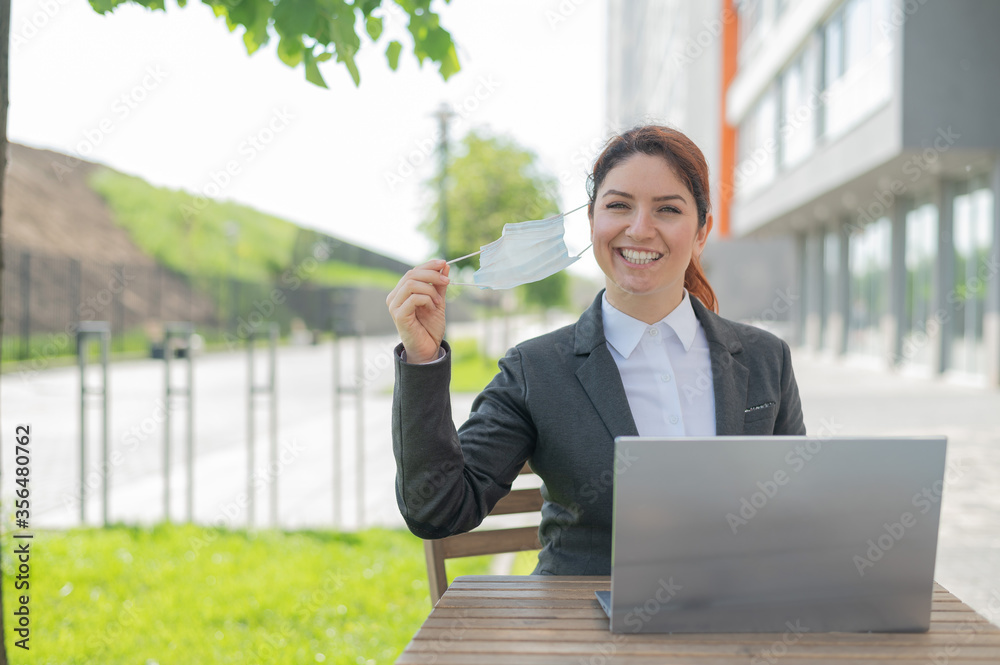 Happy business woman removes a medical mask while working on a laptop on a summer terrace. A smiling girl in a suit enjoys freedom while sitting in a cafe. End of quarantine. Happy business woman removes a medical mask while working on a laptop on a summer terrace. A smiling girl in a suit enjoys freedom while sitting in a cafe. End of quarantine.