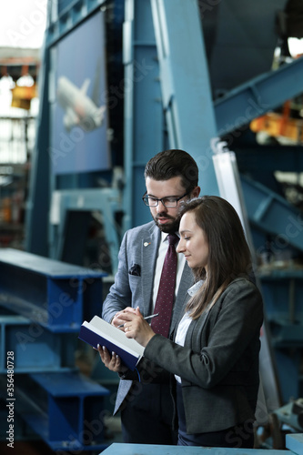 Portrait of a solid businessman with his secretary holding blue notebook, talking about factory financial report in a airplane manufactory. Factory managers controlling work process in a factory