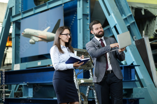  Portrait of a solid businessman with his secretary holding blue notebook, talking about factory financial report in a airplane manufactory. Factory managers controlling work process in a factory