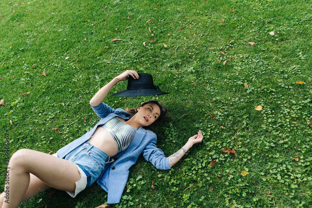 Fashion portrait of a young girl dressed in a blue striped jacket and black hat. Girl lies on a green lawn in a park in the city Fashion portrait of a young girl dressed in a blue striped jacket and black hat. Girl lies on a green lawn in a park in the city