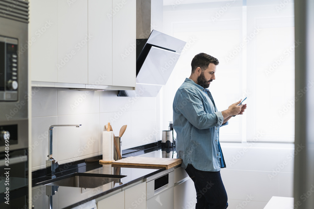 Man cooking crepes in the kitchen with a mobile phone in a denim shirt Man cooking crepes in the kitchen with a mobile phone in a denim shirt
