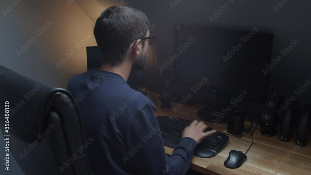 Young man sitting at his desk with two black monitors over shoulder shot Young man sitting at his desk with two black monitors over shoulder shot