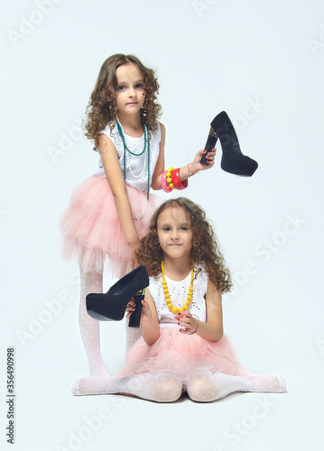 two little girls play with their mother's high-heeled shoes. they pose and try to be adults.Studio, White background