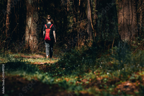 Lonely young girl with backpack goes along picturesque path into dark forest. Female teenager wandering in woods or lost. Free life of youth, independence or freedom of choice. Run away from home