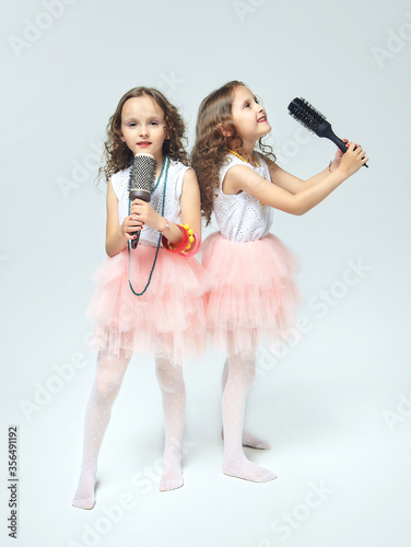 two little girls comb each other's hair. twin sisters singing into hair brushes. white background