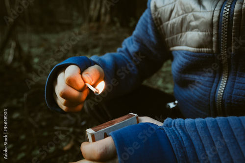 Naughty kid playing with burning matches in dark forest. Danger of burns to children. Matchbox and flame in hands of a child. Unsafe handling of fire and attention of parents. Accident or risk