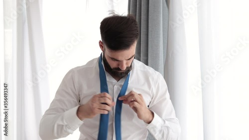 Young bearded handsome man in white shirt trying to tie his neck tie preparing for first job interview or date and having issue because it is his first time and it is complicate photo