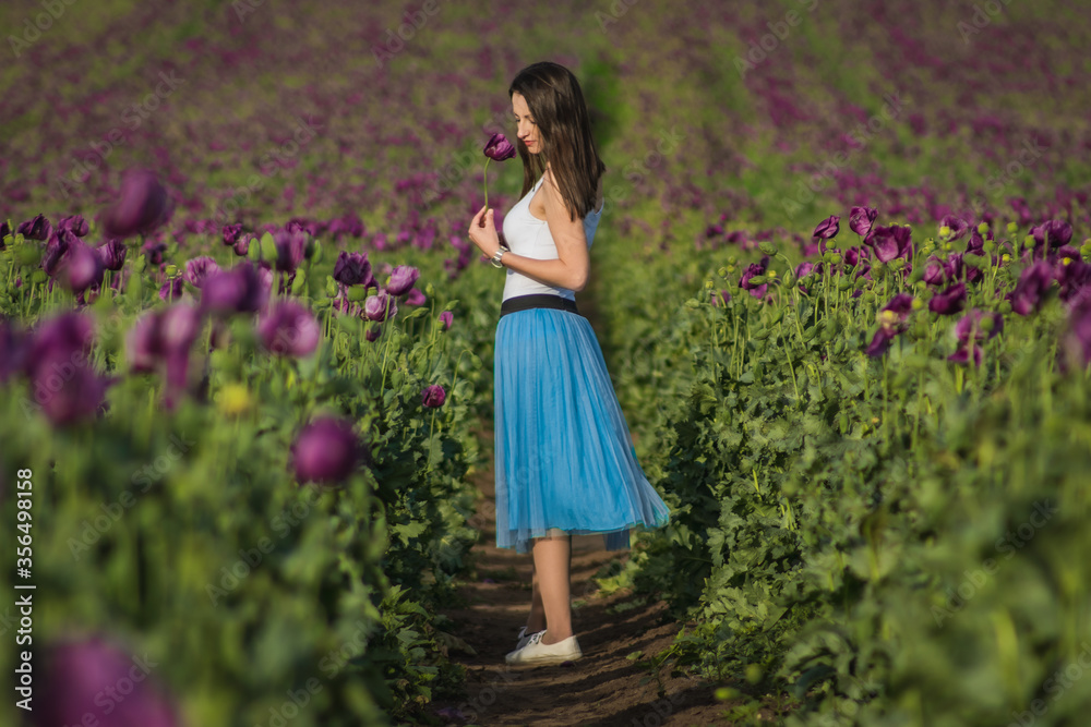 Happy beautiful woman model in a blue skirt standing on a path of the blooming summer field full of purple poppy flowers Happy beautiful woman model in a blue skirt standing on a path of the blooming summer field full of purple poppy flowers