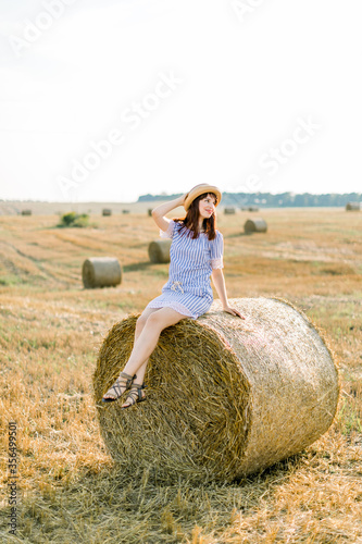 Charming red haired girl in hat and summer striped dress sitting on a hay bale in warm summer sunny day, wheat field on the background. Woman looking away and smiling