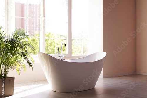 Modern bathroom. White bath with green palm branches. Bright room with sunlight from a large window © brillianata