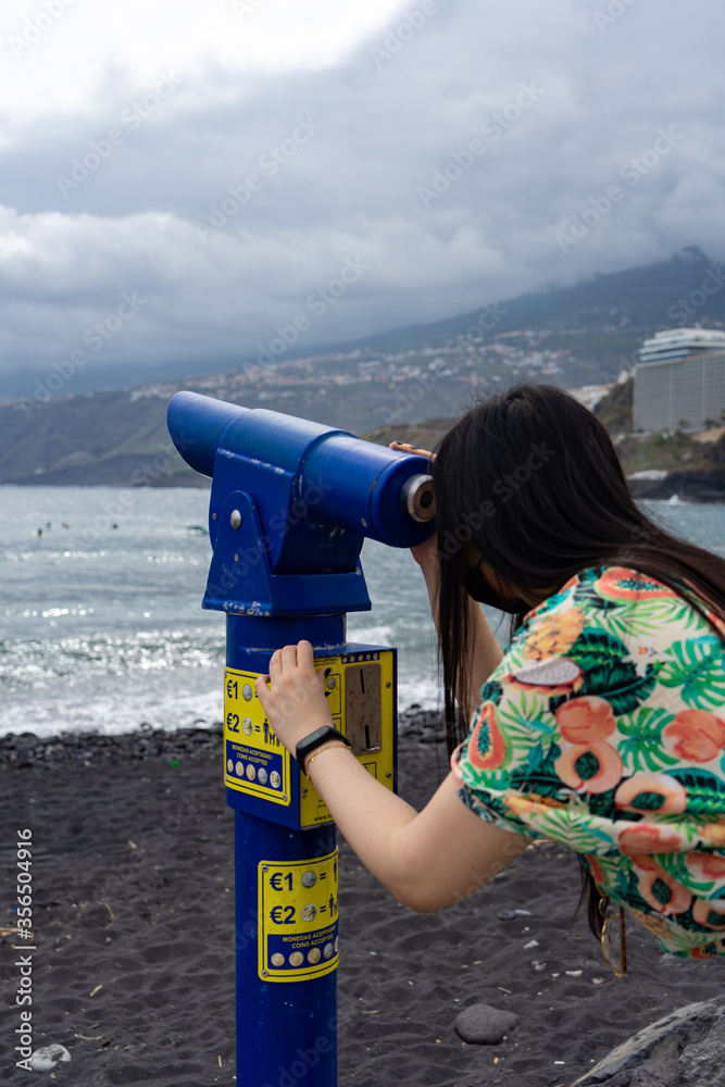 Asian woman looking through a binocular. It is located in a viewpoint of the city of Puerto De La Cruz, Tenerife. She wears a floral shirt and a mask.