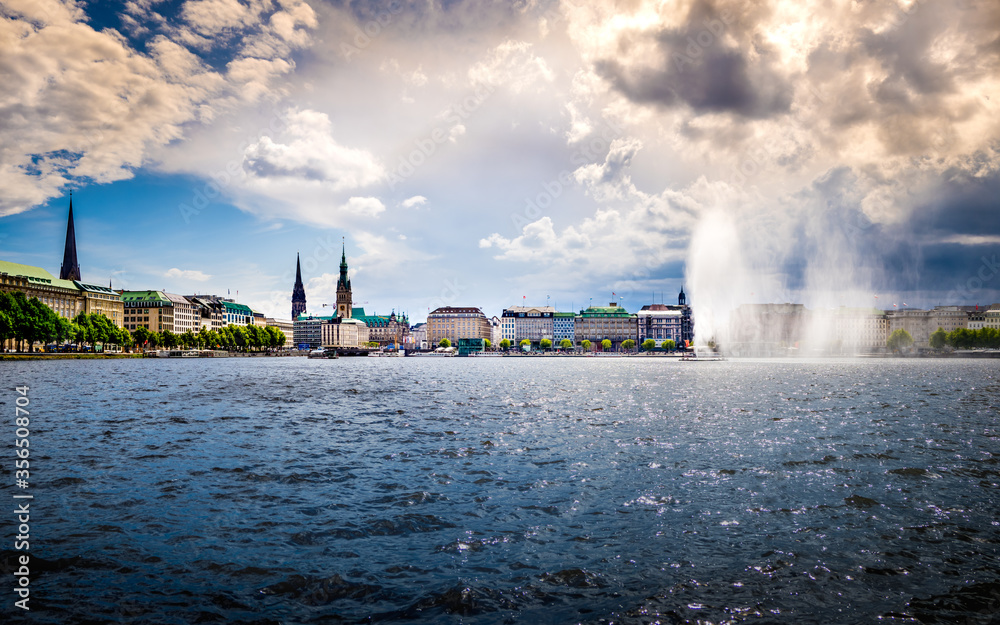 idyllic horizontal panorama of the binnenalster waterfront in hamburg old town with view to the jungfernstieg with town hall and majestic church towers in the background under dramatic cloudscape